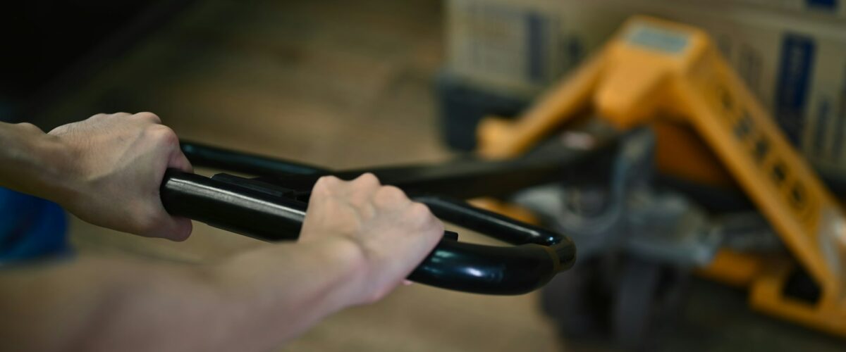 Man worker pulling hand pallet truck with cardboard package boxes in distribution warehouse.